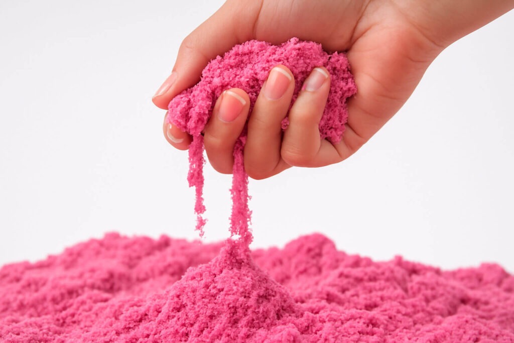 Ръка стиснала розов кинетичен пясък, който се разпада над купчина от същия пясък върху бял фон.