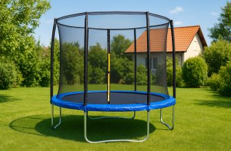 Кръгъл детски батут със син ръб и мрежа за безопасност, поставен на трева с къща и дървета в далечината.