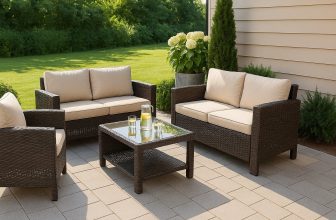 Комплект ратанови градински мебели с бежови възглавници и стъклена масичка, разположени върху плочки в озеленен двор