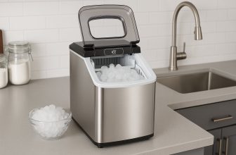 Снимка на компактна машина за лед от неръждаема стомана, поставена върху светъл кухненски плот до мивка, с купа пълна с лед отпред, в чиста и подредена кухня с бели плочки. Alt текст: Машина за лед от неръждаема стомана в кухня с купа лед до нея.