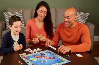 Семейство от трима души играе настолна игра Монополи на кръгла маса в уютна дневна, всички са усмихнати и ангажирани с играта.