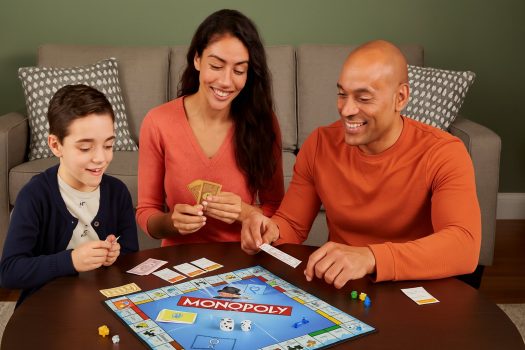 Семейство от трима души играе настолна игра Монополи на кръгла маса в уютна дневна, всички са усмихнати и ангажирани с играта.