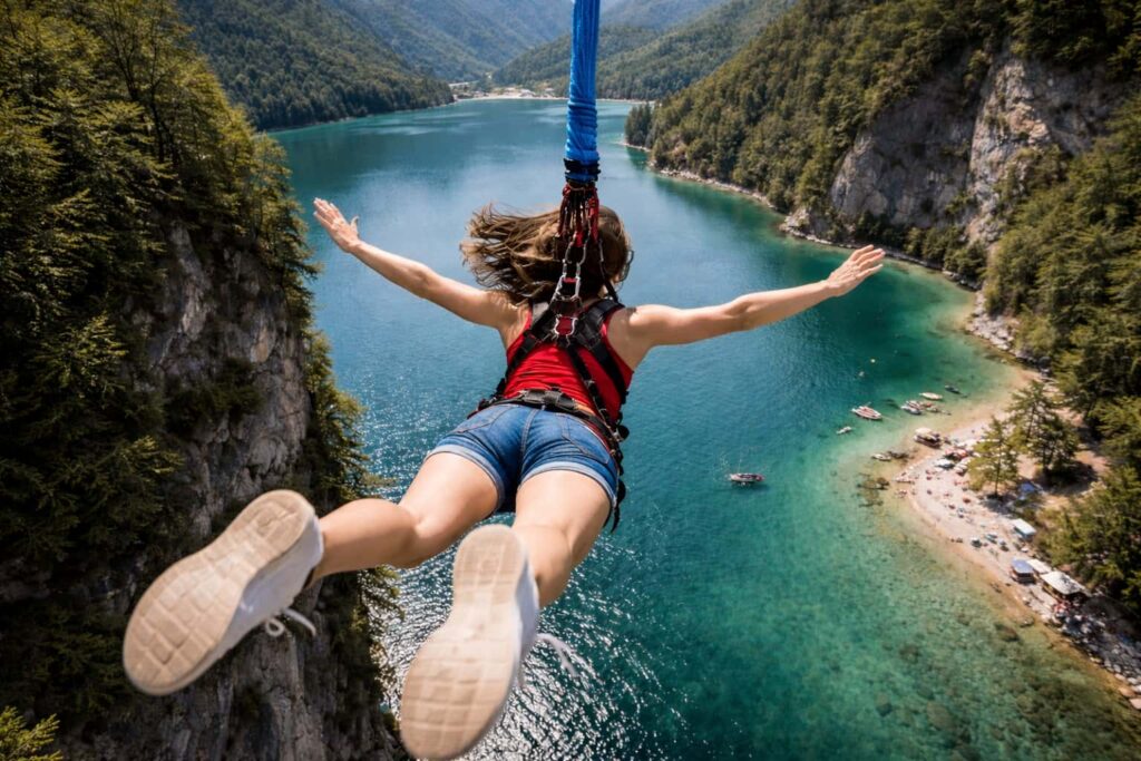 Изглед отзад на човек, който скача с бънджи над тюркоазено планинско езеро, с въже закрепено към хамут на гърба.